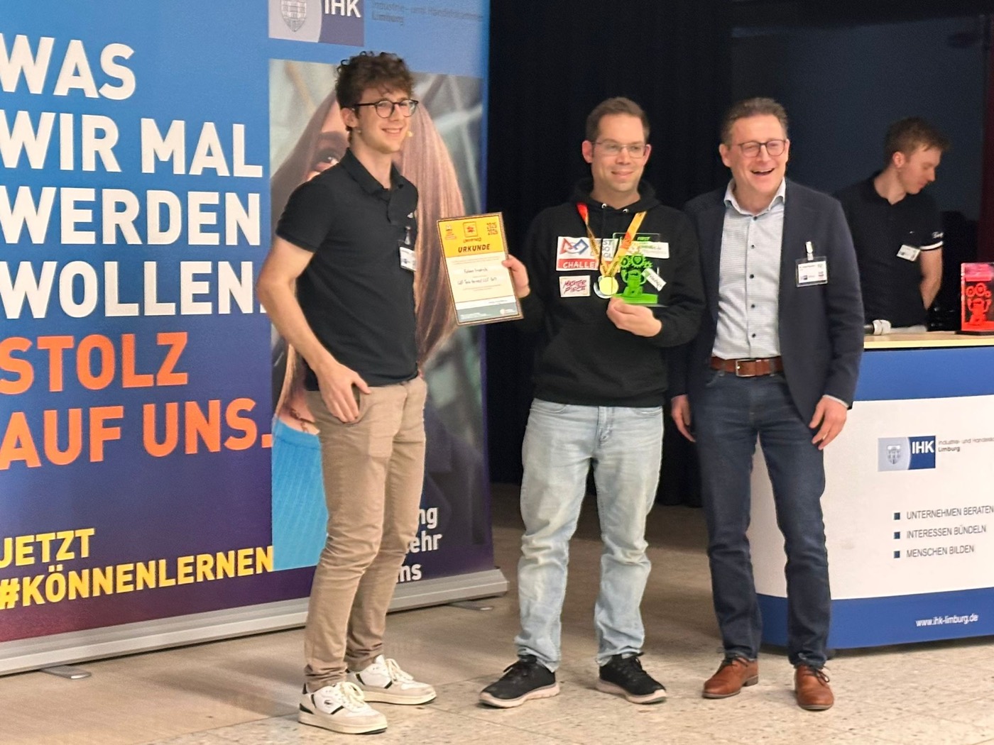 Übergabe des Coachingpokals auf der Bühne: der Betreuer der Gutenbergschule hält Urkunde und Pokal, links der Moderator, rechts ein Vertreter der IHK Limburg