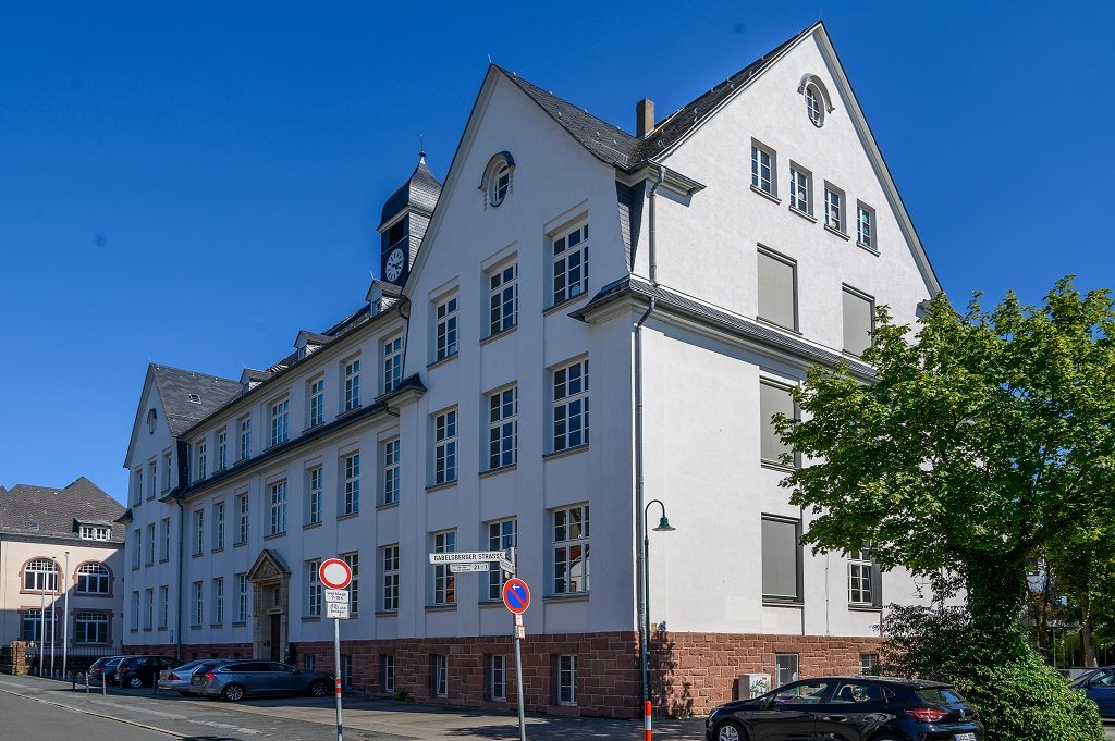 Gebäude A heute: heller Putzbau mit rotem Sandsteinsockel, Mittelturm mit Uhr, davor die Gabelsberger Straße