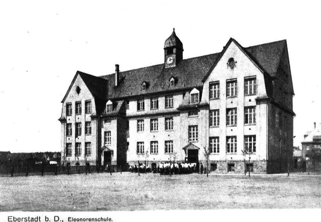 Historische Aufnahme des dreigliedrigen Schulgebäudes mit Mittelturm, Turmuhr und geschwungener Haube; vor dem Eingang steht eine Gruppe von Kindern