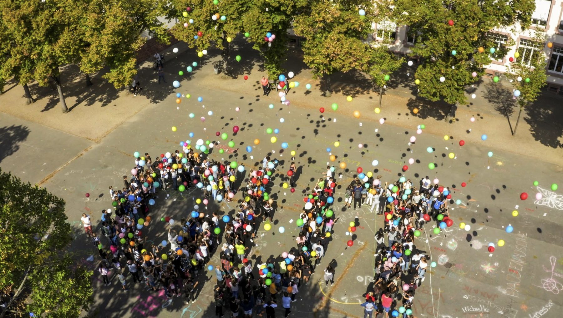 Luftaufnahme des Schulhofs: Die Schulgemeinschaft steht in Form der Buchstaben GUT zusammen, über ihr steigen Hunderte bunte Luftballons in den Himmel
